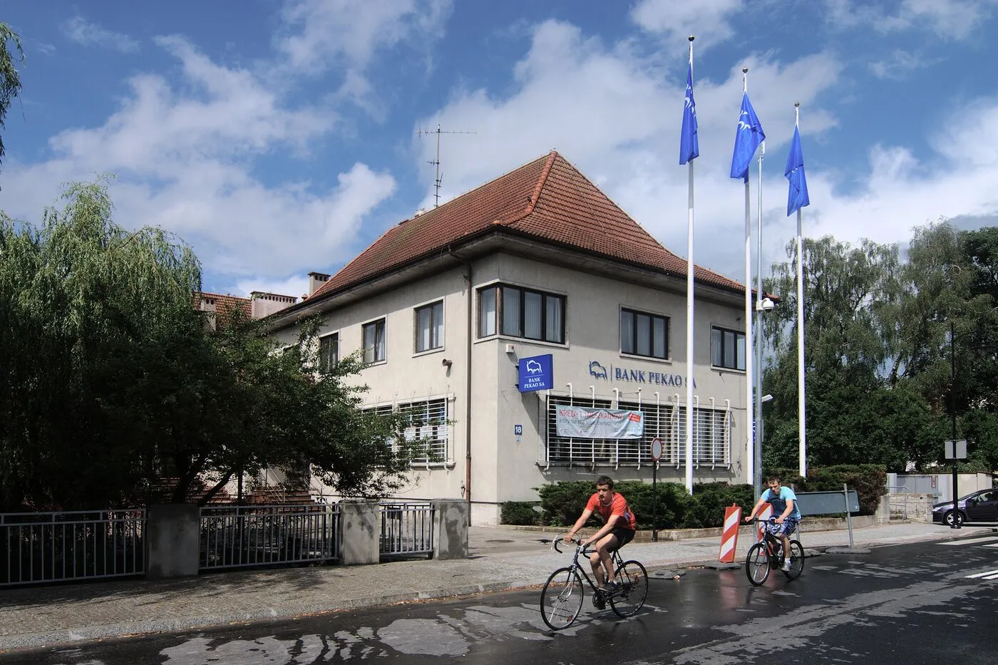 Budynek banku przy ulicy Armii Krajowej 18: niski, tynkowany, z czerwonym dachem, nad korytem rzeki.