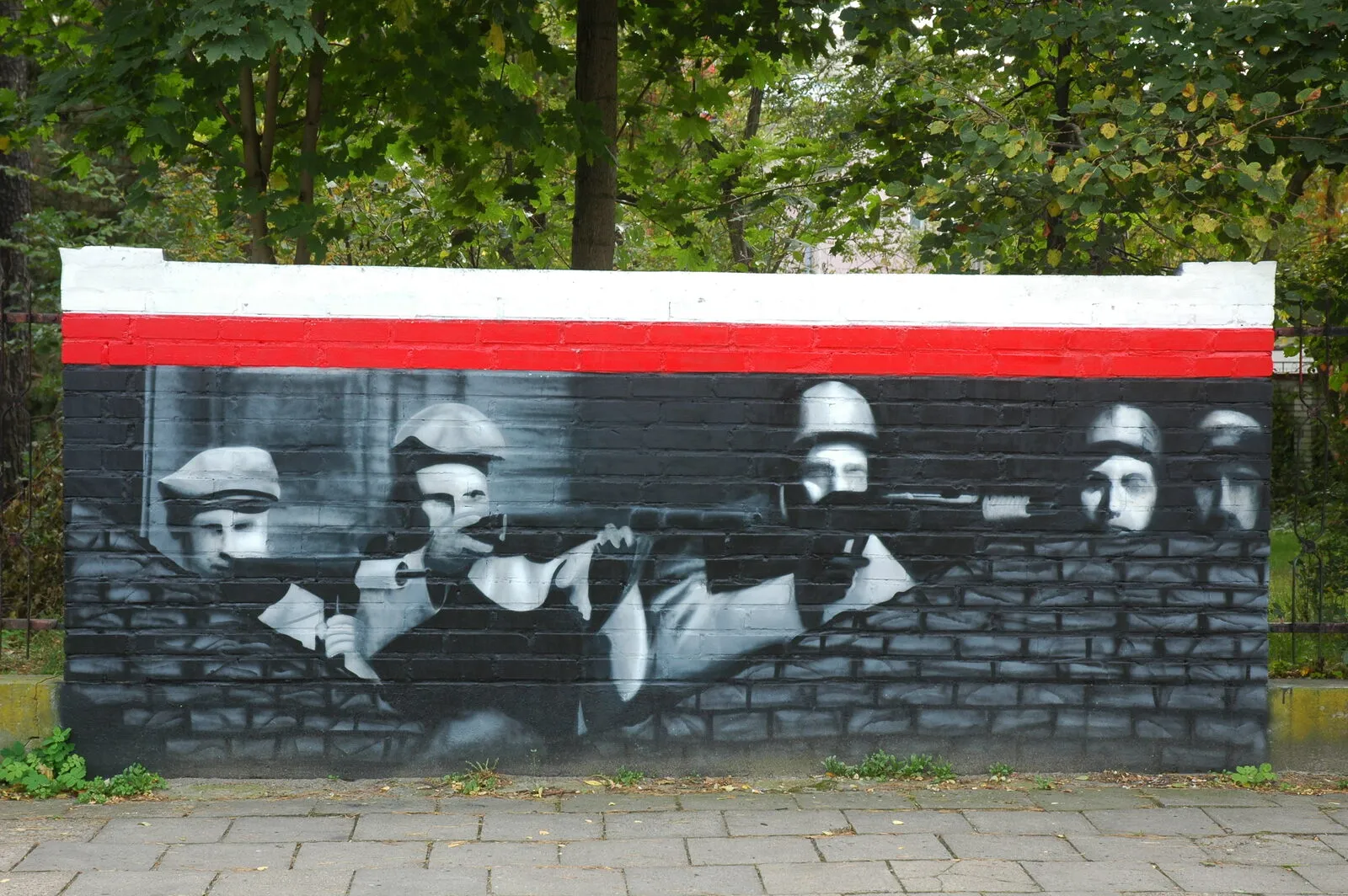 Uliczny mural na ceglanym murze: biało-czarne postacie żołnierzy w hełmach z karabinami, nad nimi poziomy biało-czerwony pas. Legionowo, róg Batorego i Siemiradzkiego.
