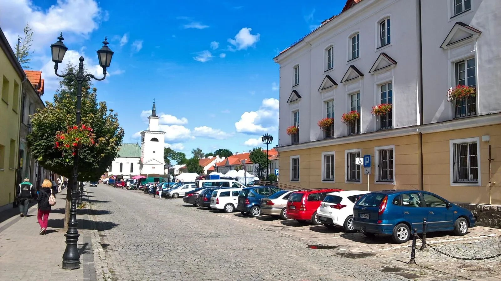 Długi brukowany rynek w Pułtusku z ceglaną wieżą ratuszową pośrodku i rzędami kamienic po bokach.