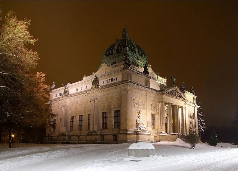 Oświetlony nocą gmach Miejskiego Domu Kultury w Zgorzelcu z kolumnowym portykiem, rzeźbami przy schodach i zieloną kopułą, przed nim śnieg.