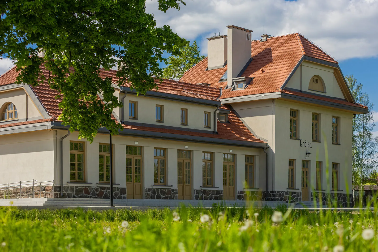 Odrestaurowany budynek dworca grójeckiej kolei wąskotorowej: kremowa elewacja na kamiennym cokole, czerwony dach z lukarnami, wysokie okna i drzwi oraz napis Grójec na ścianie szczytowej.