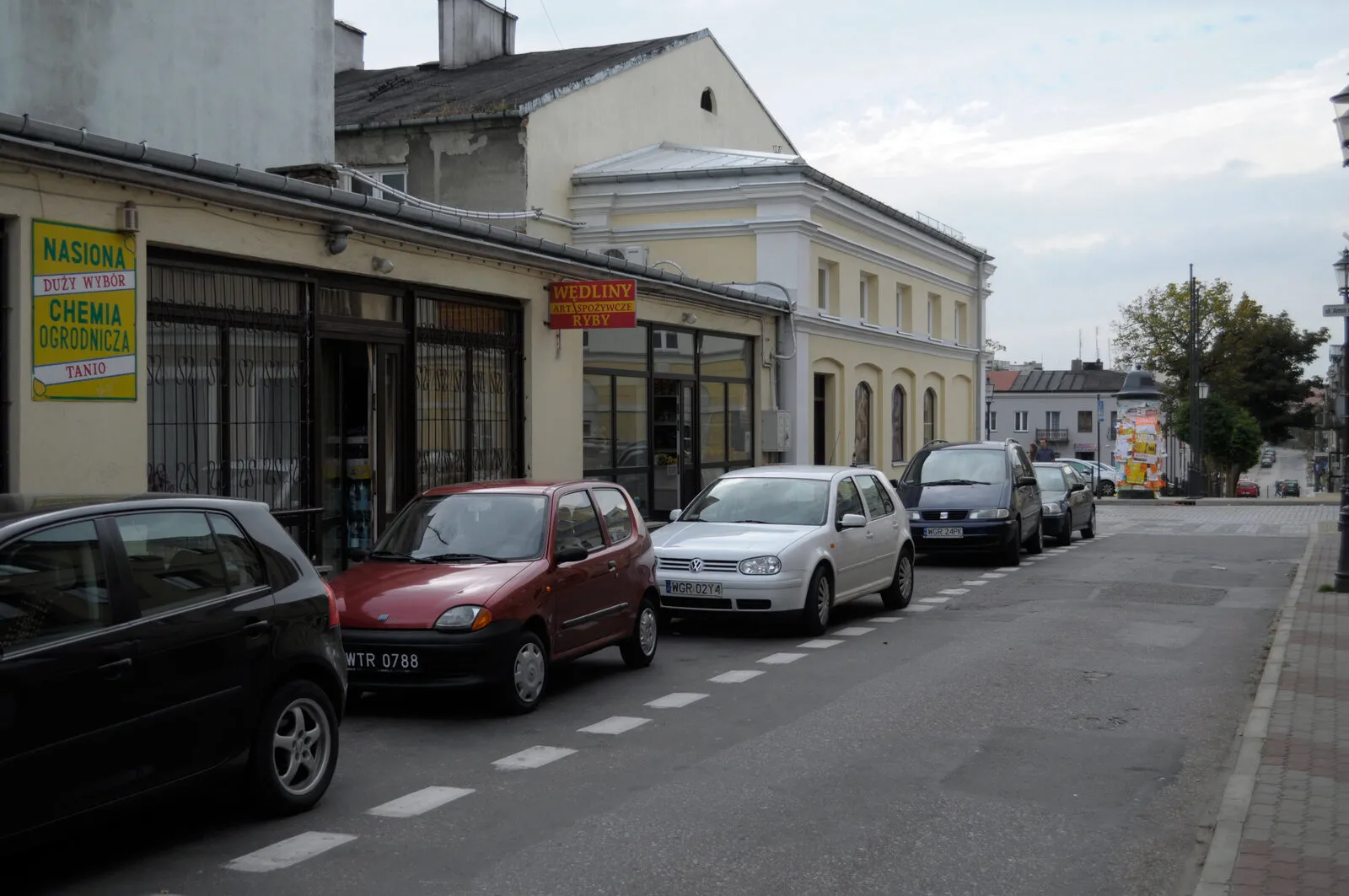 Pierzeja dawnych jatek przy ulicy Jatkowej w Grójcu: parterowe i piętrowe budynki z witrynami sklepowymi i szyldami sklepów spożywczych, wzdłuż ulicy zaparkowane samochody.
