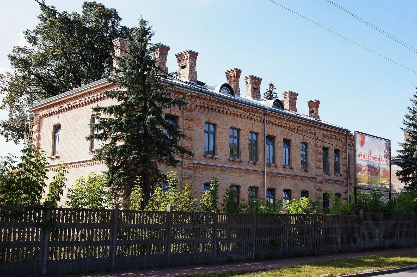 Ceglany budynek koszar z ozdobnymi kominami i okienkami w dachu.