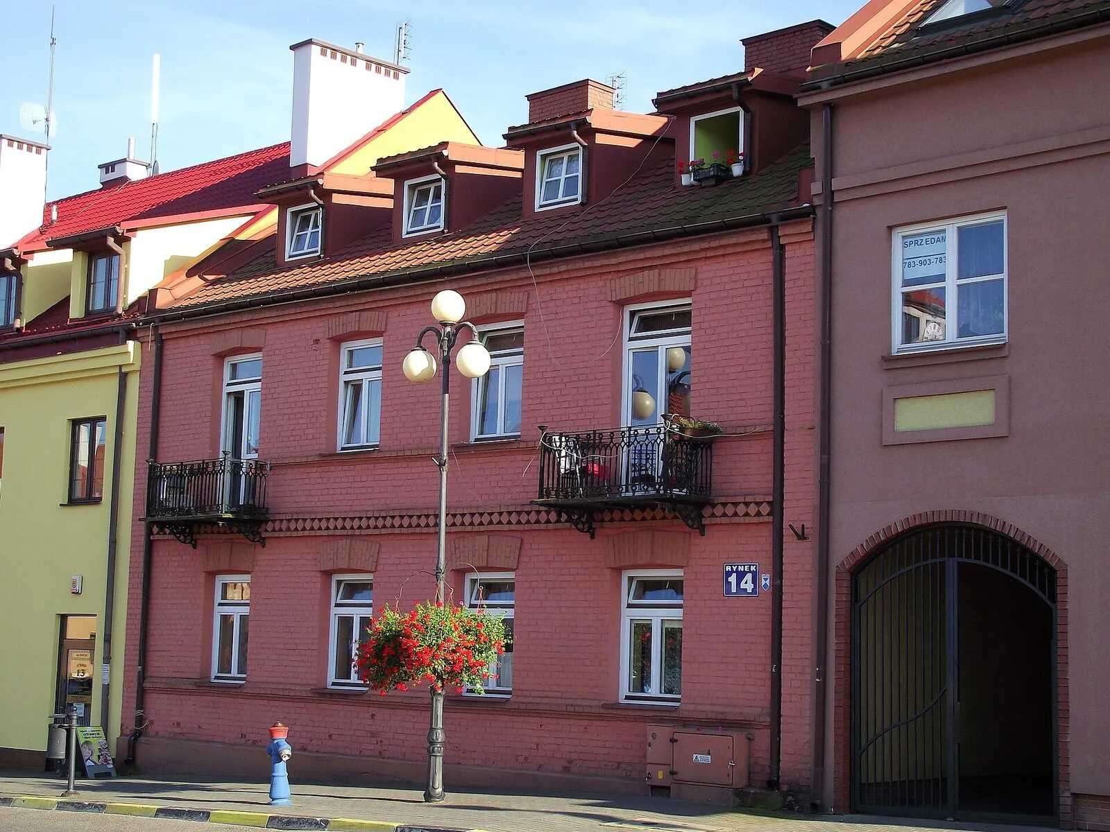 Kamienica przy Rynku 14 w Serocku z czerwonej, nieotynkowanej cegły: dwie kondygnacje, żeliwne balustrady balkonów, ceglany fryz i tabliczka z numerem 14, obok sklepiony przejazd bramny zamknięty kratą.