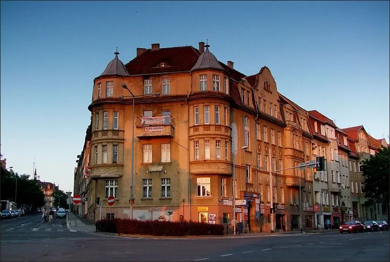 Narożna kamienica przy ulicy Warszawskiej 1 w Zgorzelcu z wykuszami, balkonami i spiczastymi hełmami wieżyczek, oświetlona wieczornym słońcem.