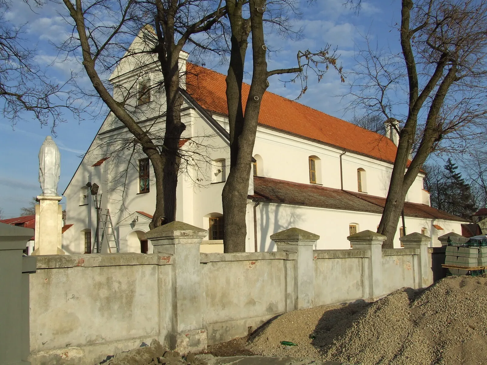 Kościół Świętej Trójcy w Błoniu, otynkowana bryła z czerwonym dachem za murowanym ogrodzeniem