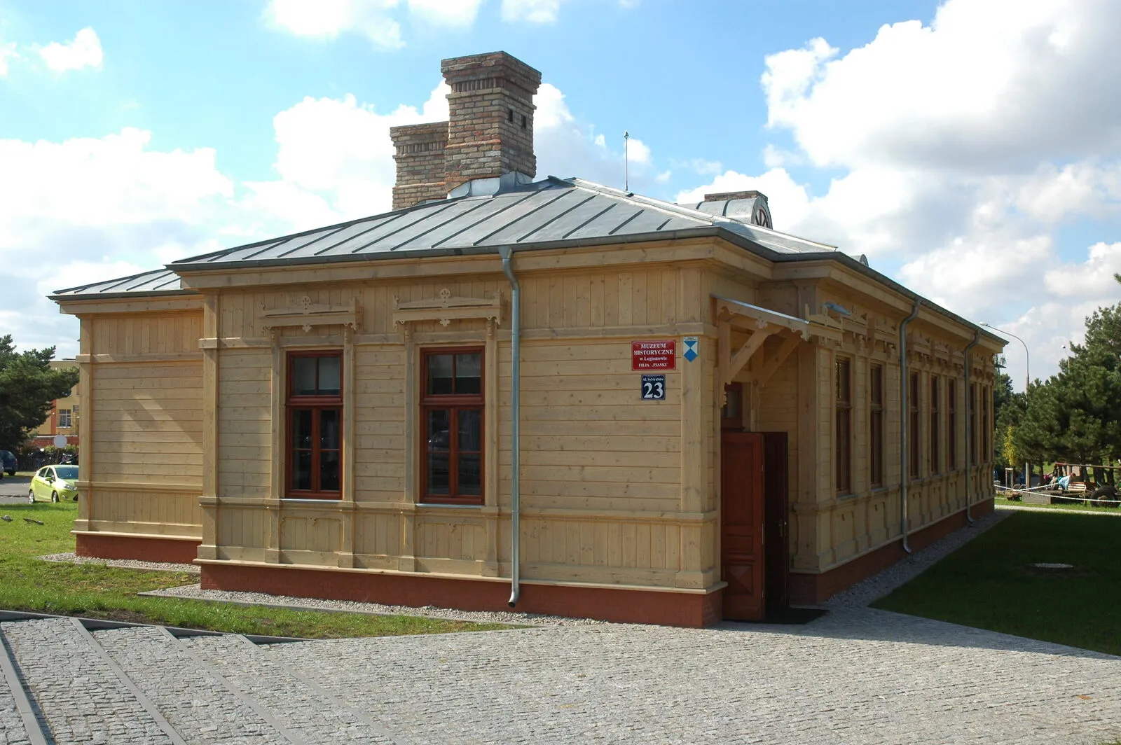 Parterowy drewniany budynek o jasnych deskowanych ścianach, z ozdobnymi nadokiennikami, wysokim ceglanym kominem i metalowym dachem. Tablica: Muzeum Historyczne w Legionowie Filia Piaski, Aleja Sybiraków 23.