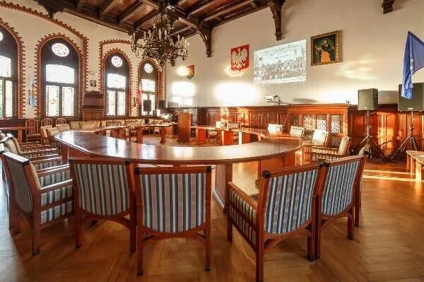 Sala Rajców: wnętrze z drewnianym stropem z belek, okrągłym stołem i oknami z łukami.