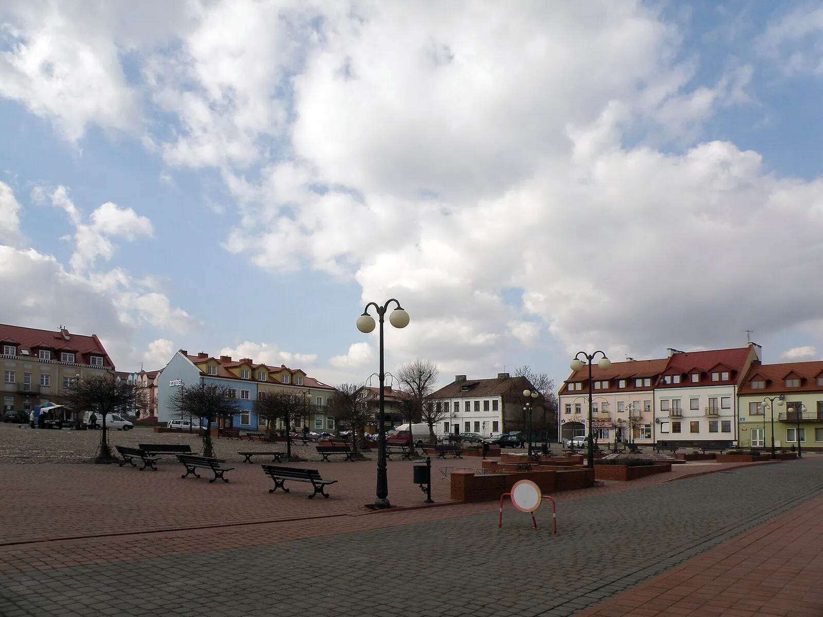 Rynek w Serocku: rozległy, brukowany plac z ławkami i stylową latarnią, wokół niska, kolorowa zabudowa z czerwonymi dachami, w tle bezlistne drzewa i zachmurzone niebo.