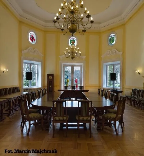 Reprezentacyjna sala na planie ośmioboku z długim stołem, żyrandolem i trzema oknami.
