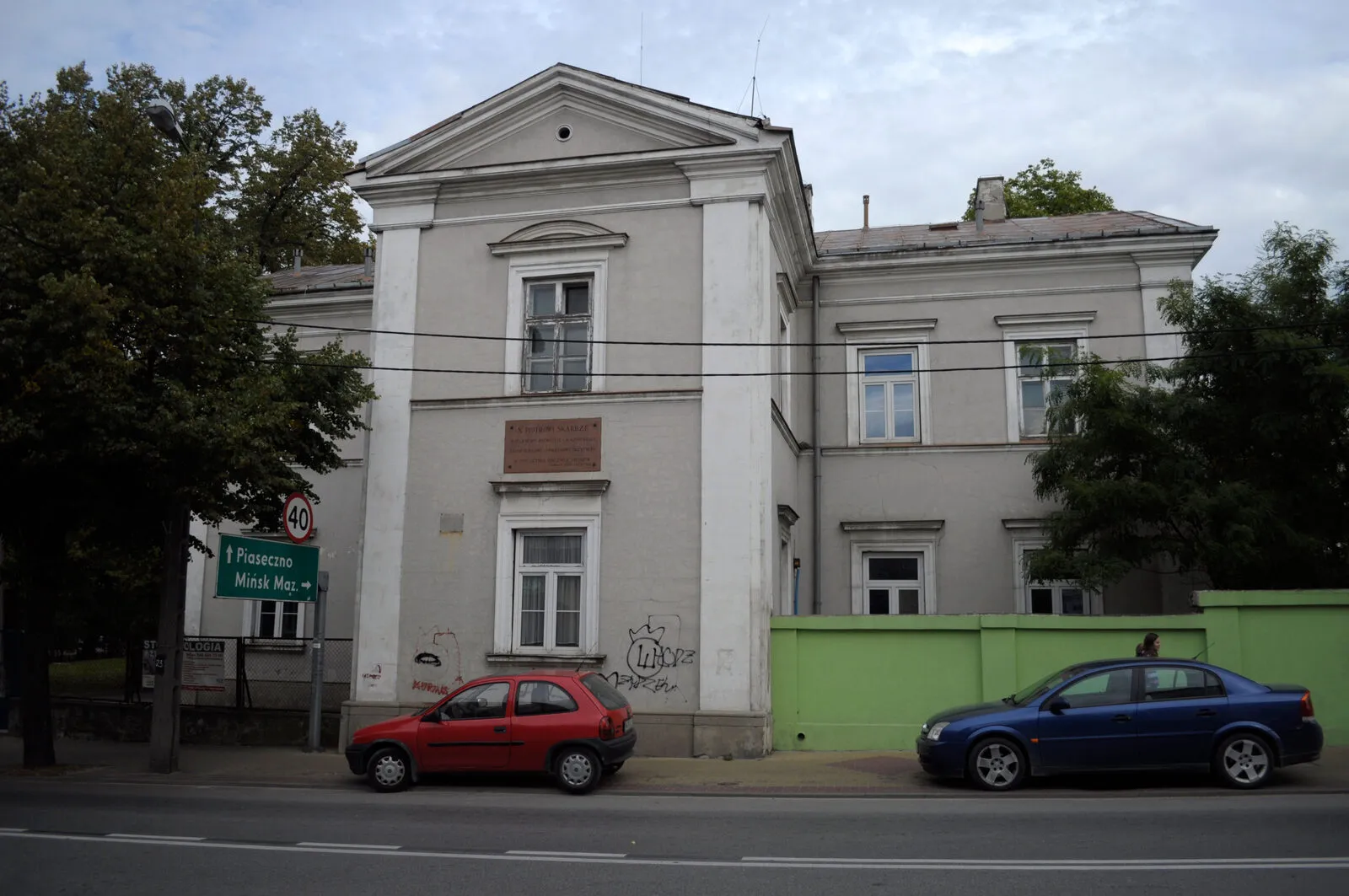 Klasycystyczny budynek dawnego szpitala w Grójcu: szara fasada z trójkątnym szczytem i pilastrami, na ścianie tablica poświęcona księdzu Piotrowi Skardze.